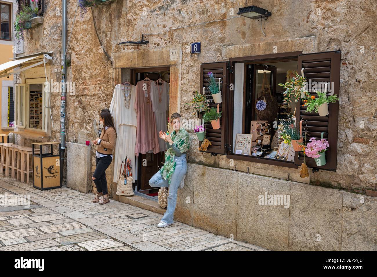 Porec, Croatie - 21 mai 2025 : deux jeunes femmes debout devant une petite boutique vendant des vêtements et des accessoires faits à la main dans une charmante rue pavée Banque D'Images
