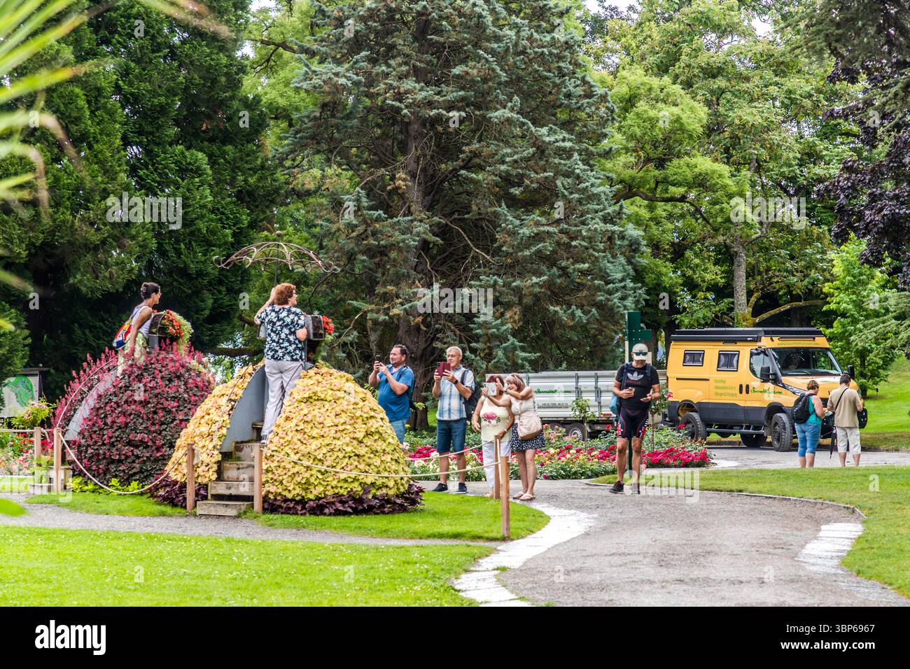 Les visiteurs profitent d'une journée dans le parc fleuri sur l'île de Mainau à Constance. Ils admirent les arrangements floraux artistiques, y compris les grands monticules fleuris, et prennent des photos sur des piédestaux de fleurs. Une camionnette de travail jaune appartenant à l'équipe d'accrobranche est garée en arrière-plan Banque D'Images Les visiteurs profitent d'une journée dans le parc fleuri sur l'île de Mainau à Constance. Ils admirent les arrangements floraux artistiques, y compris les grands monticules fleuris, et prennent des photos sur des piédestaux de fleurs. Une camionnette de travail jaune appartenant à l'équipe d'accrobranche est garée en arrière-plan Banque D'Images