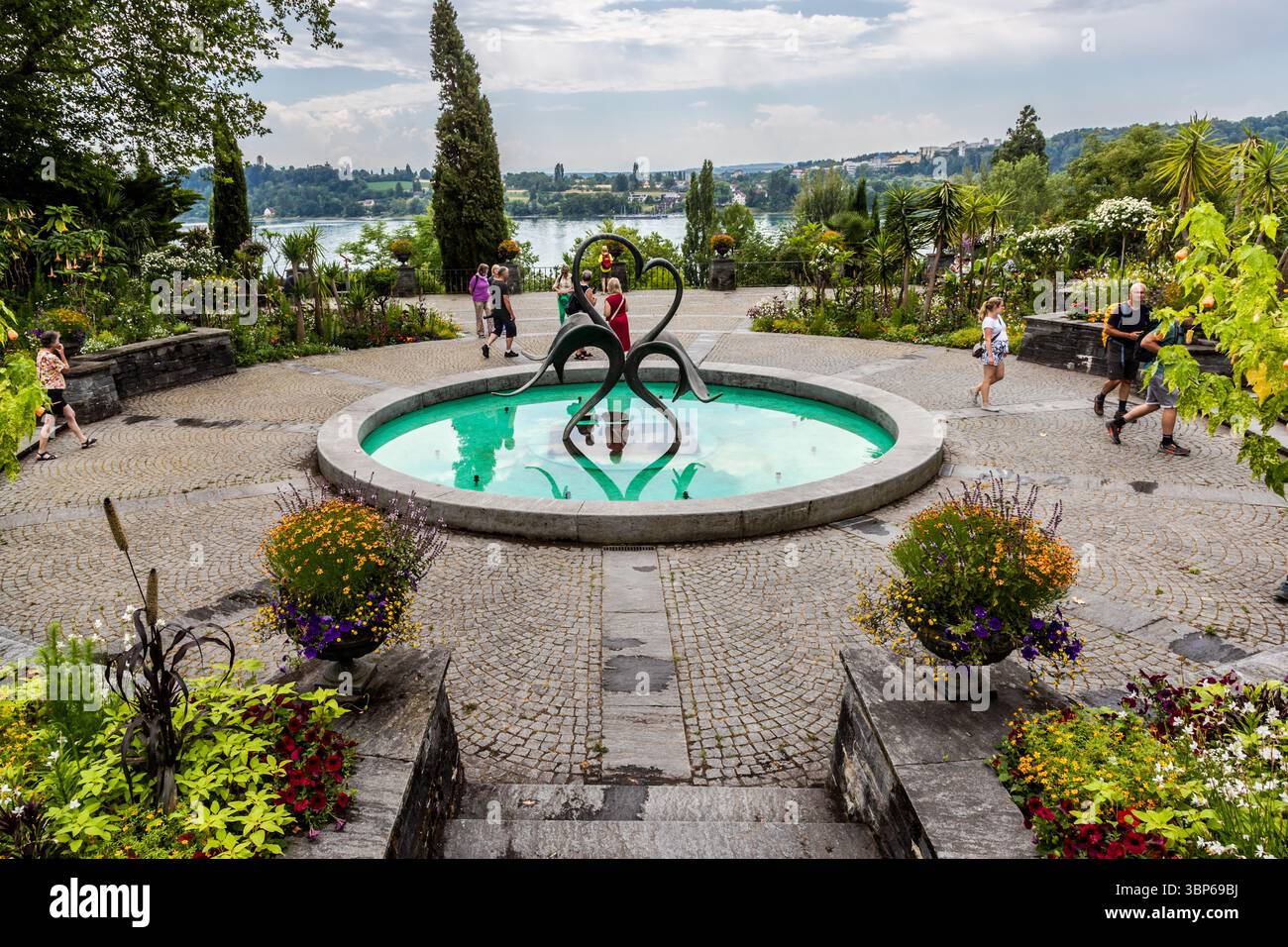 Les visiteurs se promènent dans les magnifiques jardins de l'île aux fleurs de Mainau à Constance, en Allemagne. Au centre de l'arène des fontaines se trouve une fontaine ronde ornée d'une sculpture moderne, entourée de parterres de fleurs colorées et d'une végétation luxuriante. Le lac de Constance peut être vu en arrière-plan avec le paysage riverain en face, créant une atmosphère idyllique Banque D'Images Les visiteurs se promènent dans les magnifiques jardins de l'île aux fleurs de Mainau à Constance, en Allemagne. Au centre de l'arène des fontaines se trouve une fontaine ronde ornée d'une sculpture moderne, entourée de parterres de fleurs colorées et d'une végétation luxuriante. Le lac de Constance peut être vu en arrière-plan avec le paysage riverain en face, créant une atmosphère idyllique Banque D'Images