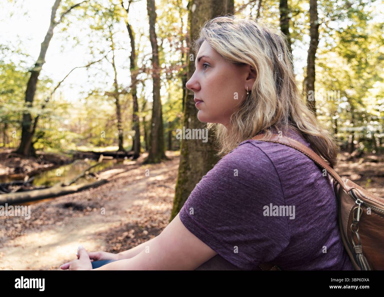 Une femme profite d'un moment où elle est assise seule sur un sentier forestier, entourée de grands arbres et baignée de lumière du soleil. Le silence permet des pensées profondes Banque D'Images