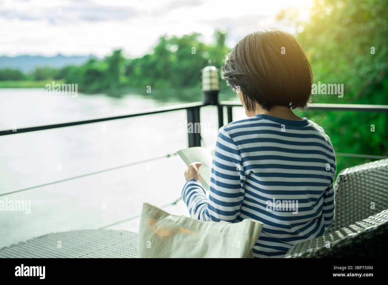 Femme assise sur la chaise lisant un livre sur le balcon surplombant la rivière pendant l'heure dorée matin. Soins personnels, détox numérique et vie lente consciente. EMO Banque D'Images