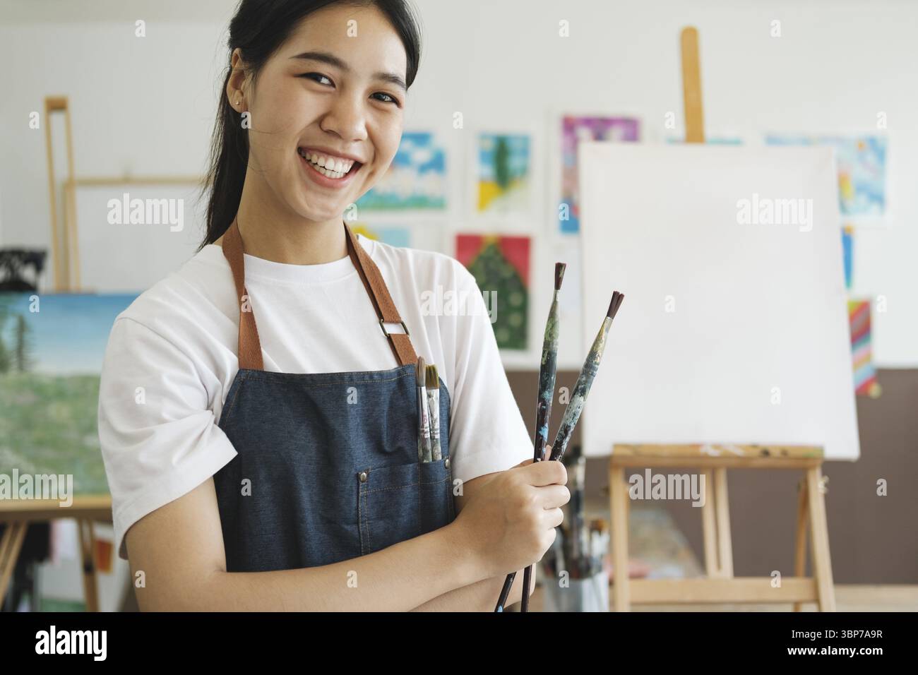 La jeune femme artiste porte un tablier enduit de peinture serrant de nombreux pinceaux et pinceaux stagnant et souriant à la caméra dans son stui Banque D'Images
