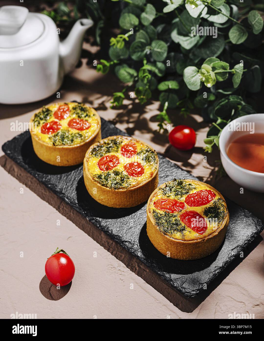 Nourriture, trois délicieuses tartes salées remplies d'épinards et surmontées de tomates cerises sont affichées sur une assiette d'ardoise. Une tasse de thé et cerise fraîche t Banque D'Images
