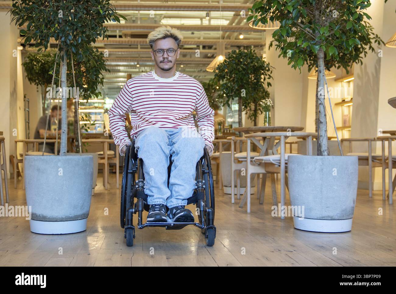 Homme handicapé confiant détendu en fauteuil roulant quittant le café, assis entre deux arbres en pot, face à la caméra. L’espace public accessible encourage l’activité Banque D'Images