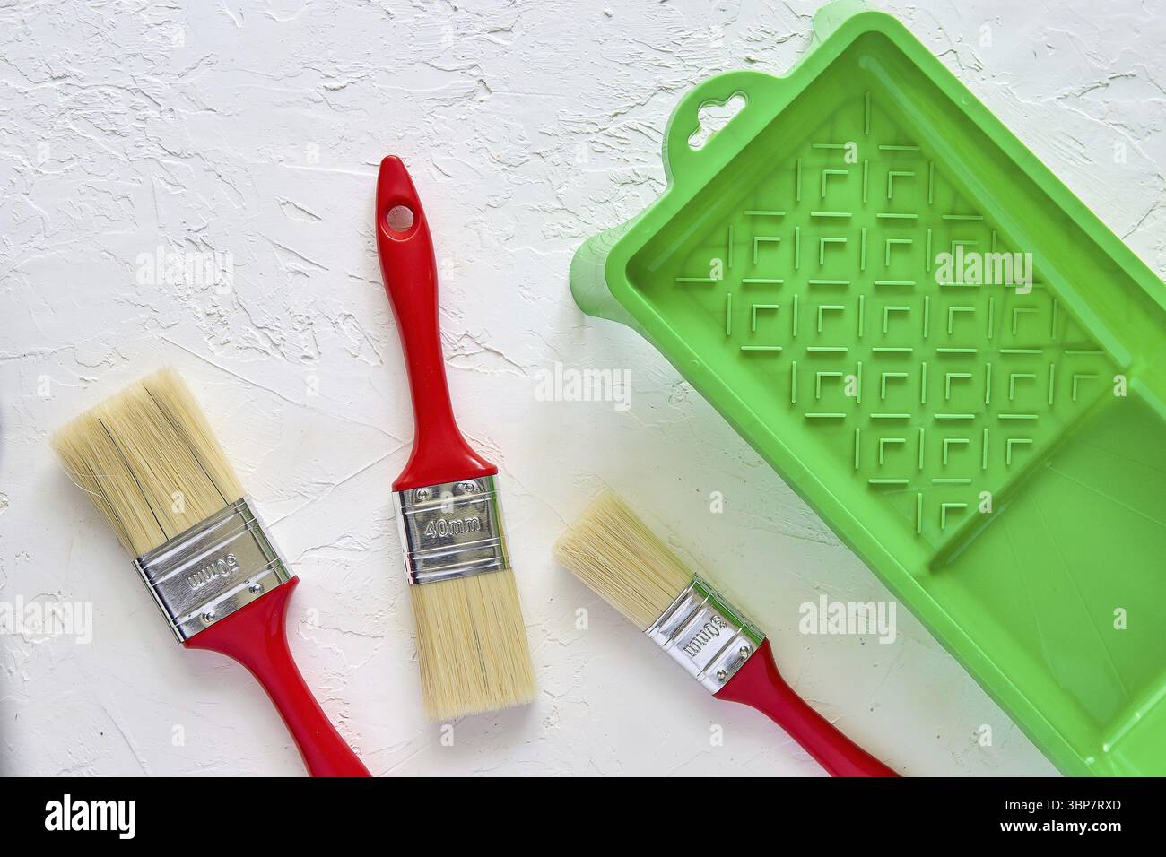 Trois brosses avec poignées rouges et bac à peinture vert sur fond de béton blanc. Outils et accessoires pour la rénovation de la maison. Vue de dessus Banque D'Images