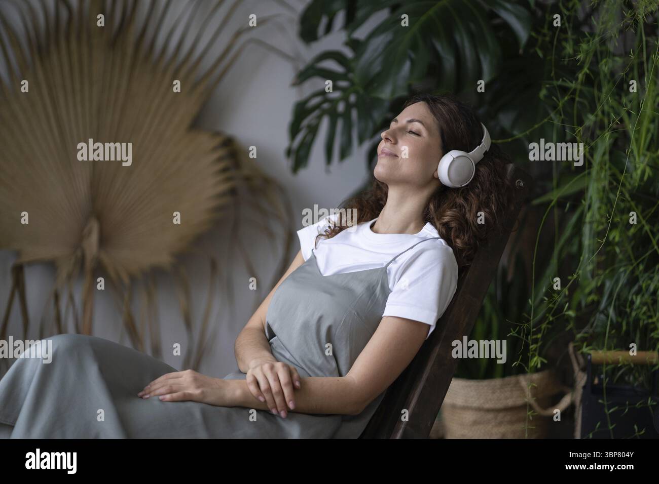 Jeune femme italienne heureuse avec les yeux fermés portant des écouteurs sans fil assis, écoutant de la musique pour réduire le stress dans la serre intérieure. La santé mentale Banque D'Images