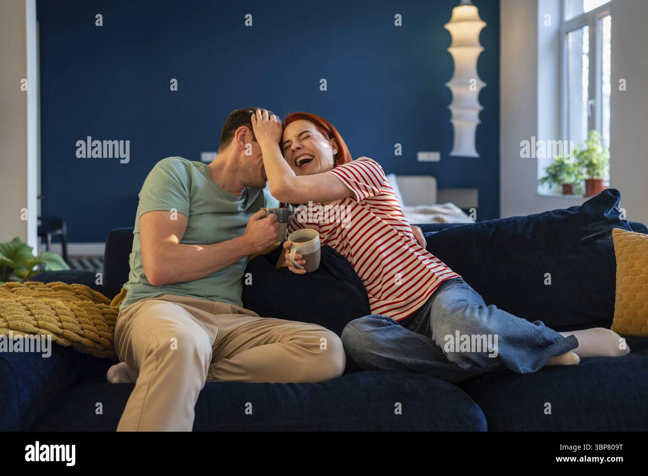 Heureux couple marié assis sur le canapé riant ensemble. Femme et homme ravi de s'amuser à la maison avec une tasse de thé. Femme souriante profiter du temps de week-end w Banque D'Images