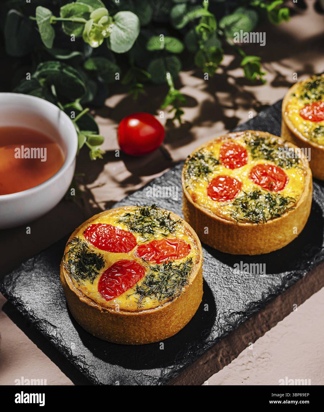 Nourriture, trois délicieuses tartes salées remplies d'épinards et surmontées de tomates cerises sont affichées sur une assiette d'ardoise. Une tasse de thé et cerise fraîche t Banque D'Images