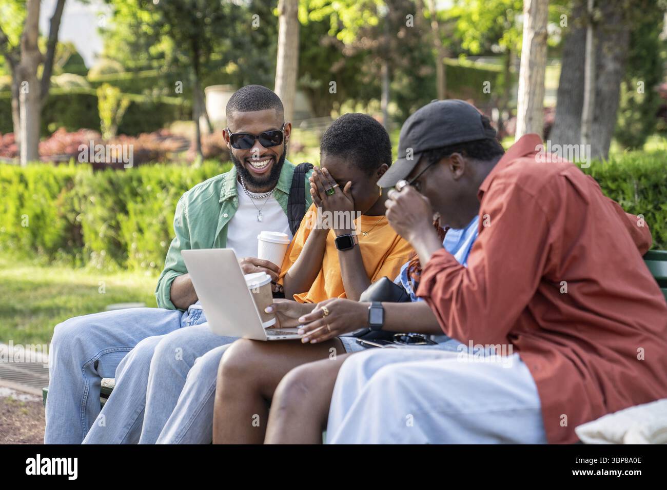 Les filles afro-américaines et les gars étudiants assises dans le parc avec un ordinateur portable riant ensemble regardant des vidéos. Heureux quatre amis noirs de diversité h Banque D'Images
