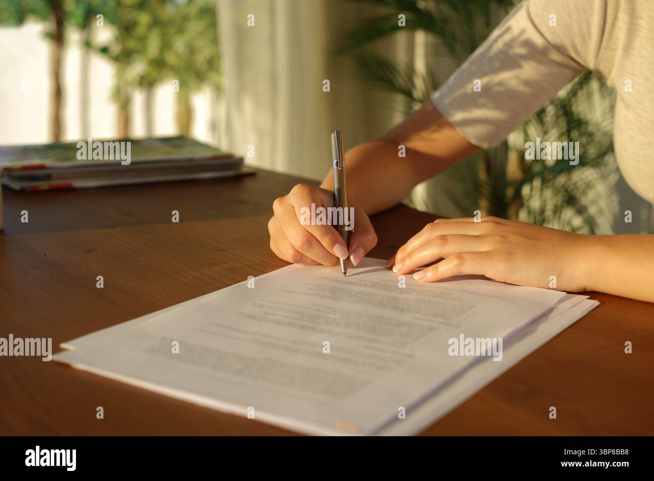 Gros plan d'une main de femme signant le document sur une table en bois à la maison au coucher du soleil Banque D'Images