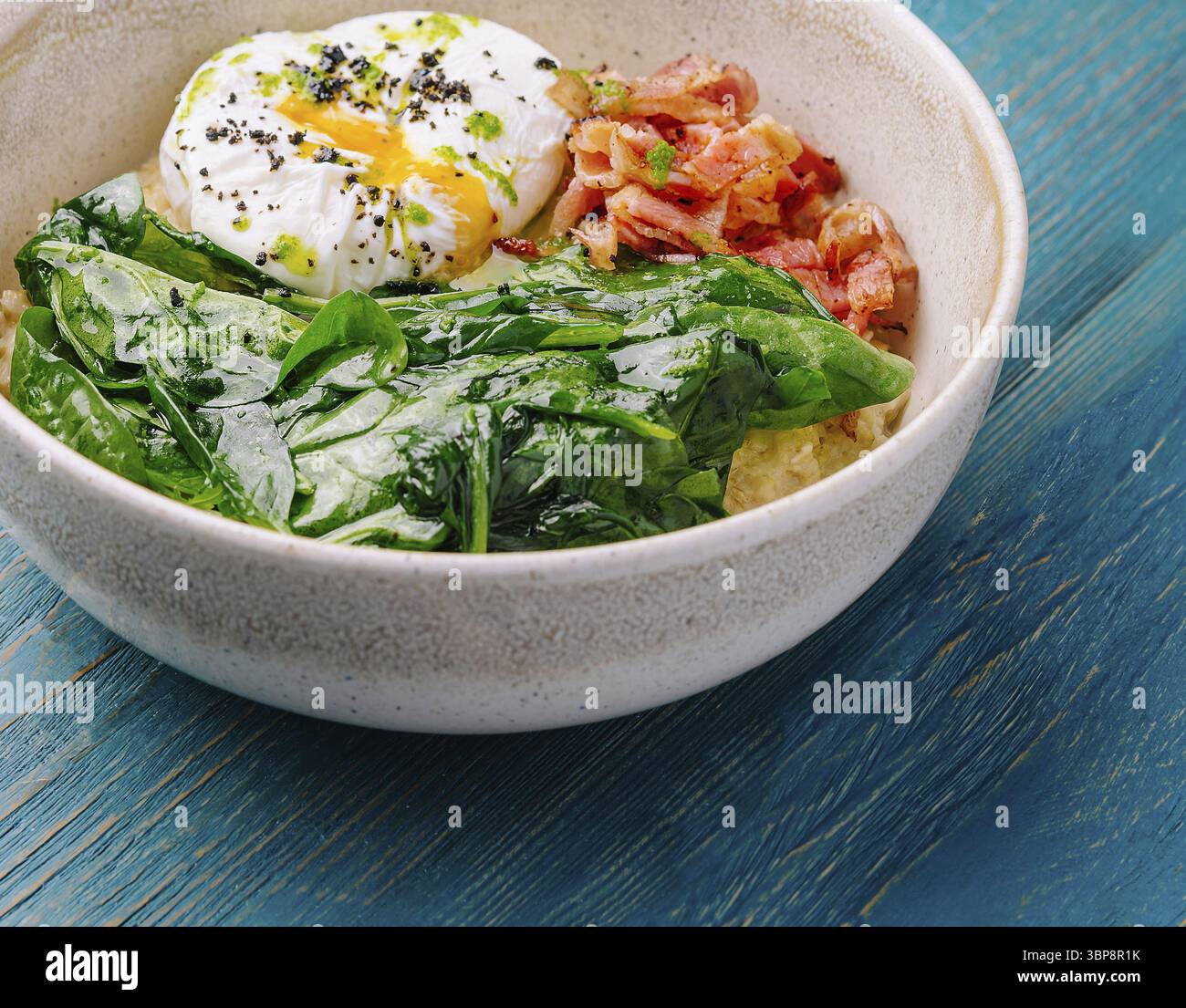 Nourriture, bol de petit déjeuner nutritif avec un œuf poché, des légumes verts fanés et du bacon croustillant sur une table bleue rustique Banque D'Images