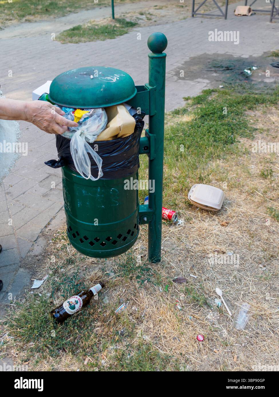 Bologne, Italie 3 juillet 2025 femme aînée jetant des ordures dans une poubelle publique débordante dans un parc à Bologne, Italie, pendant une journée d'été, avec un Banque D'Images