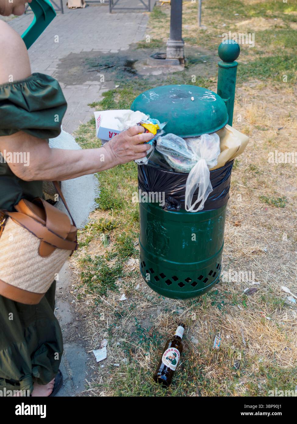 Bologne, Italie 3 juillet 2025 femme aînée jetant des ordures dans une poubelle publique débordante dans un parc à Bologne, Italie, pendant une journée d'été, avec un Banque D'Images