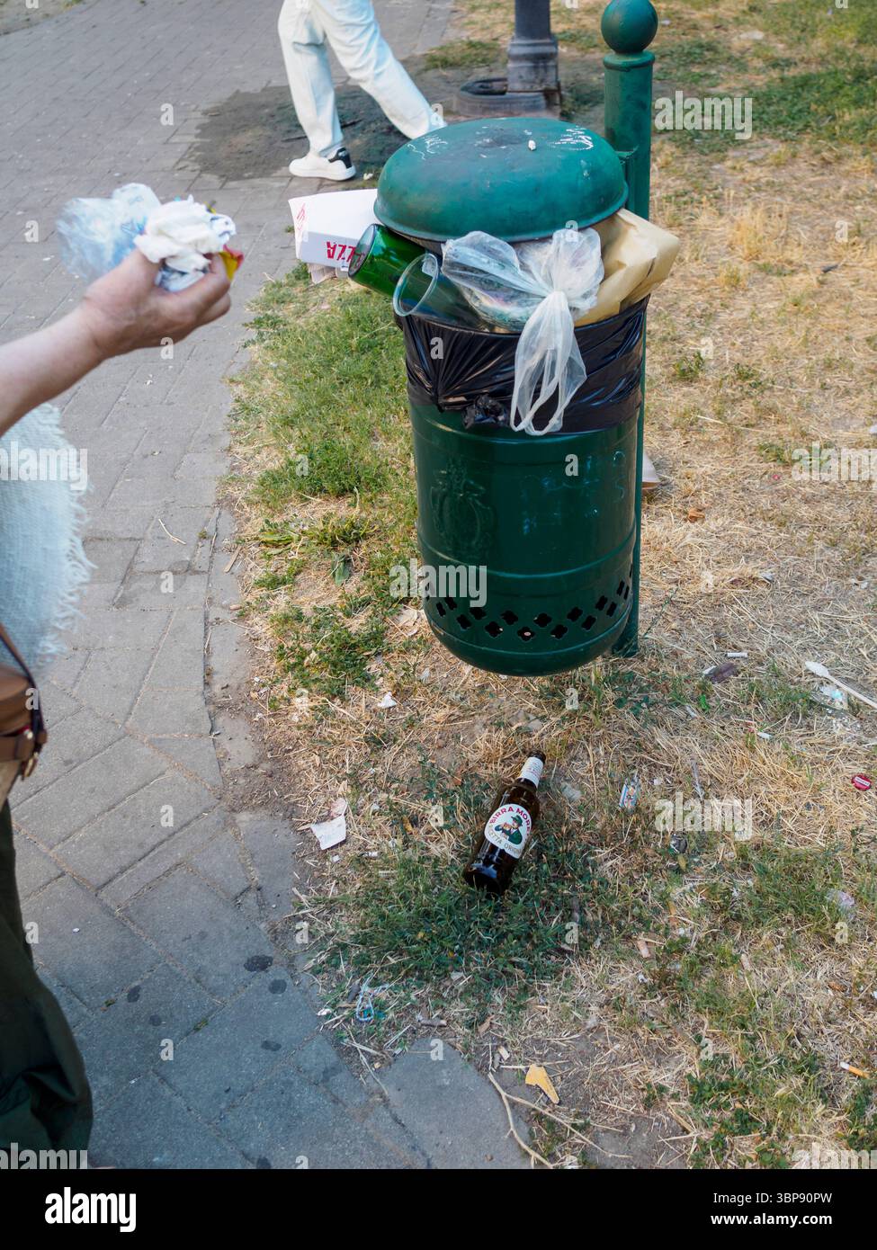 Bologne, Italie 3 juillet 2025 piéton passant le bac à ordures public trop rempli dans la rue urbaine, révélant les défis de la gestion des déchets pendant les périodes de pointe Banque D'Images