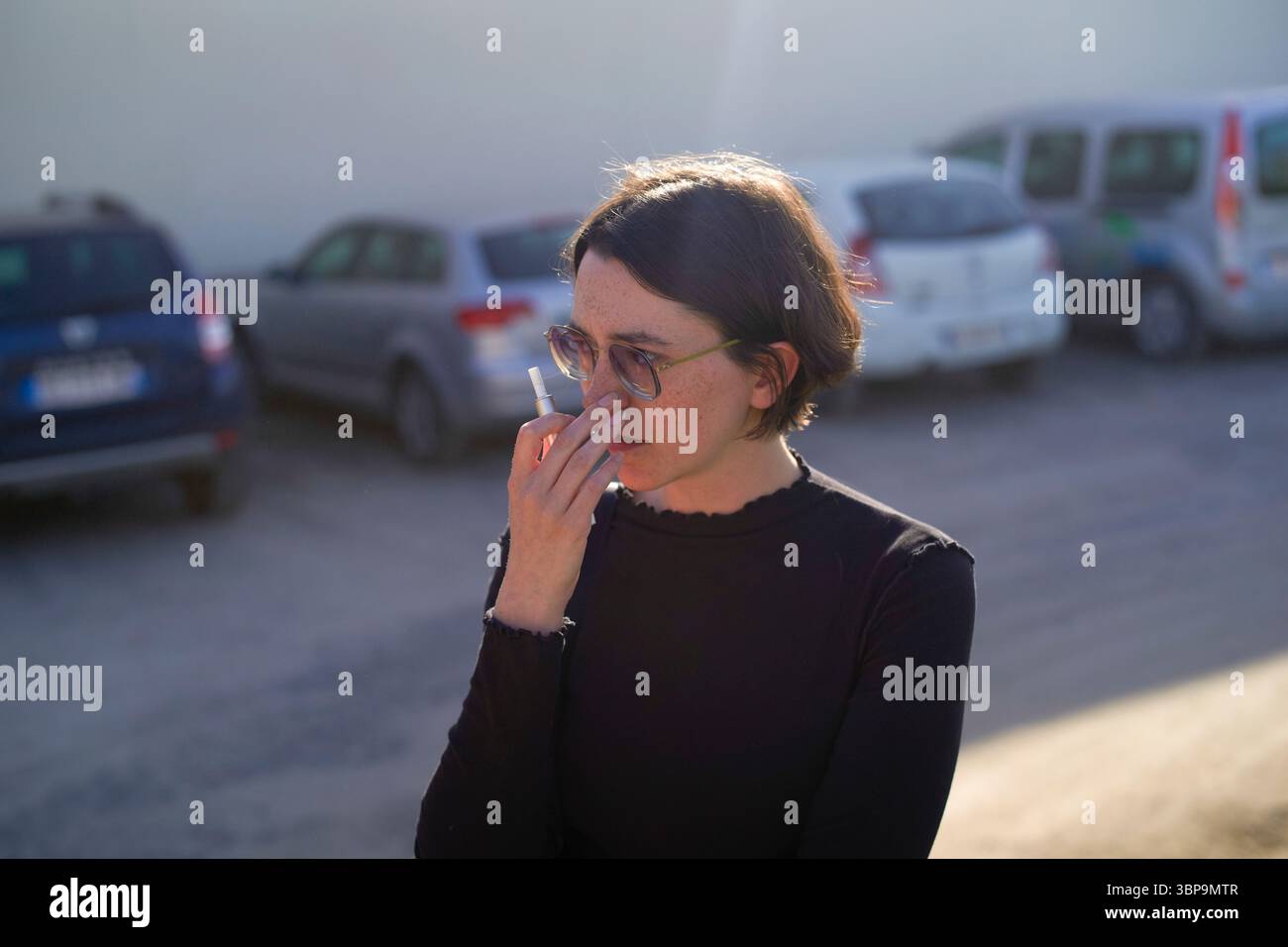 Femme avec les cheveux courts debout à l'extérieur fumant dans un parking avec des voitures garées derrière. Banque D'Images