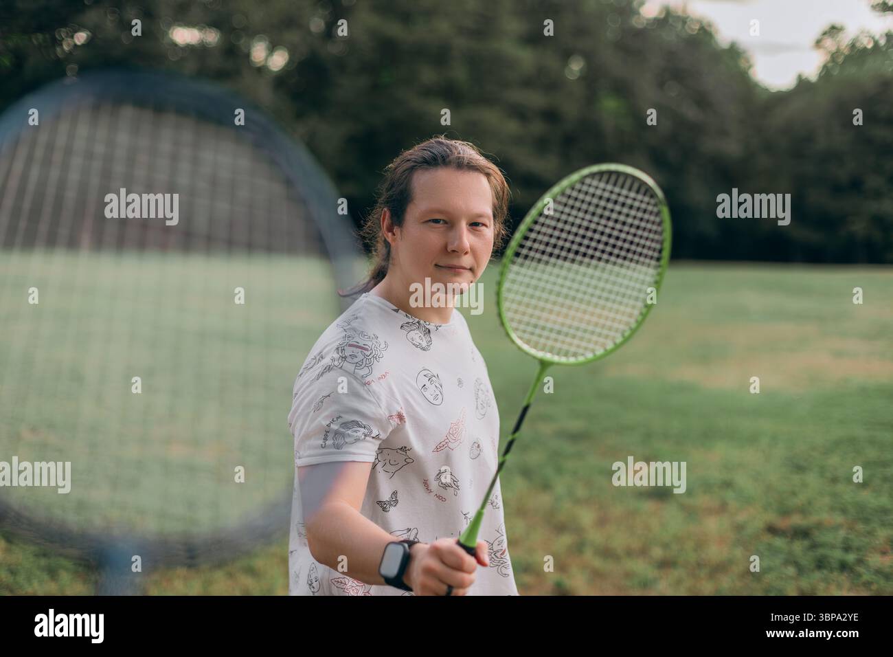 Homme souriant montrant vert badminton raquette herbeuse terrain décontracté t-shirt imprimé noir shorts smartwatch cheveux noués lumière du jour chaude Banque D'Images