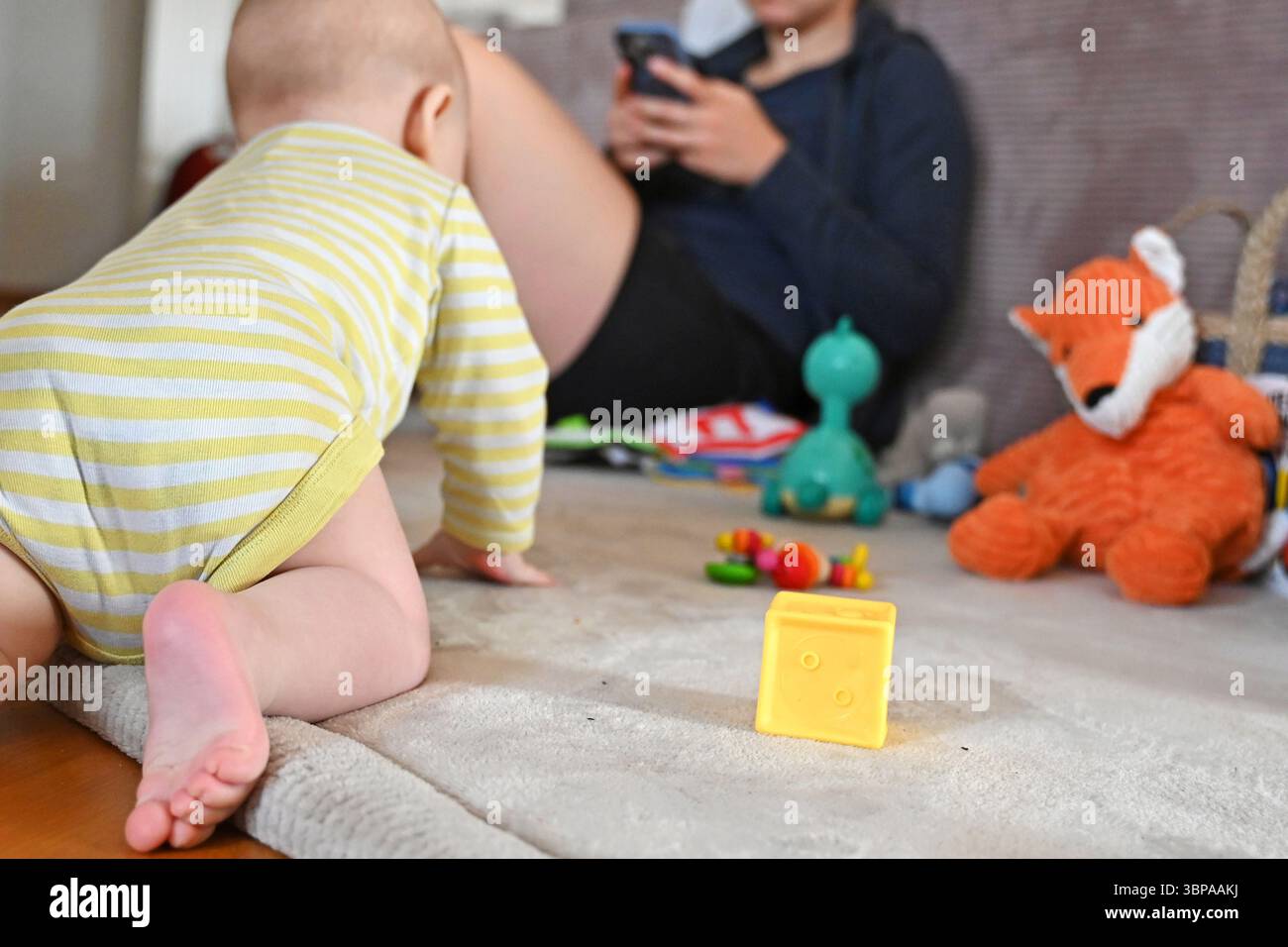 Leipzig, Allemagne. 02 juin 2025. Un bébé rampant sur un tapis vers un jouet tandis qu'une mère regarde son smartphone en arrière-plan. La vie quotidienne avec de jeunes enfants pose souvent aux parents le défi de combiner la garde des enfants avec leurs propres besoins. Crédit : ELISA Schu/dpa/Alamy Live News Banque D'Images