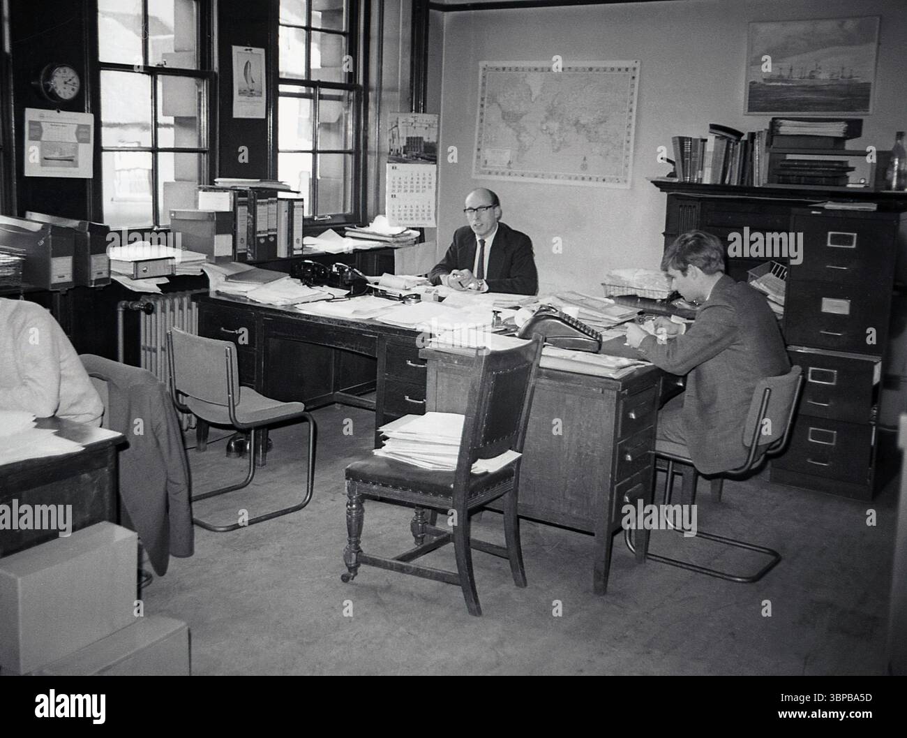1967, historique, deux employés masculins assis à des bureaux attachés dans un bureau municipal d'une compagnie maritime, Édimbourg, Écosse, Royaume-Uni. Les bureaux et les rebords des fenêtres sont recouverts de documents et de dossiers. Une carte du monde sur le mur derrière un bureau. Banque D'Images