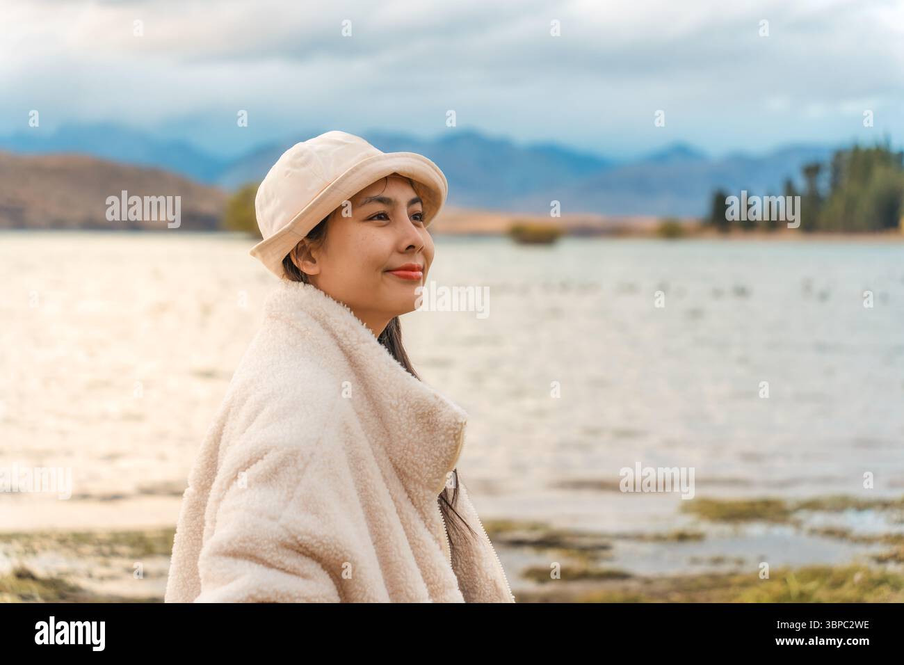 Jeune femme asiatique en veste confortable avec chapeau souriant et appréciant dans le parc d'automne par le lac Tekapo en Nouvelle-Zélande Banque D'Images