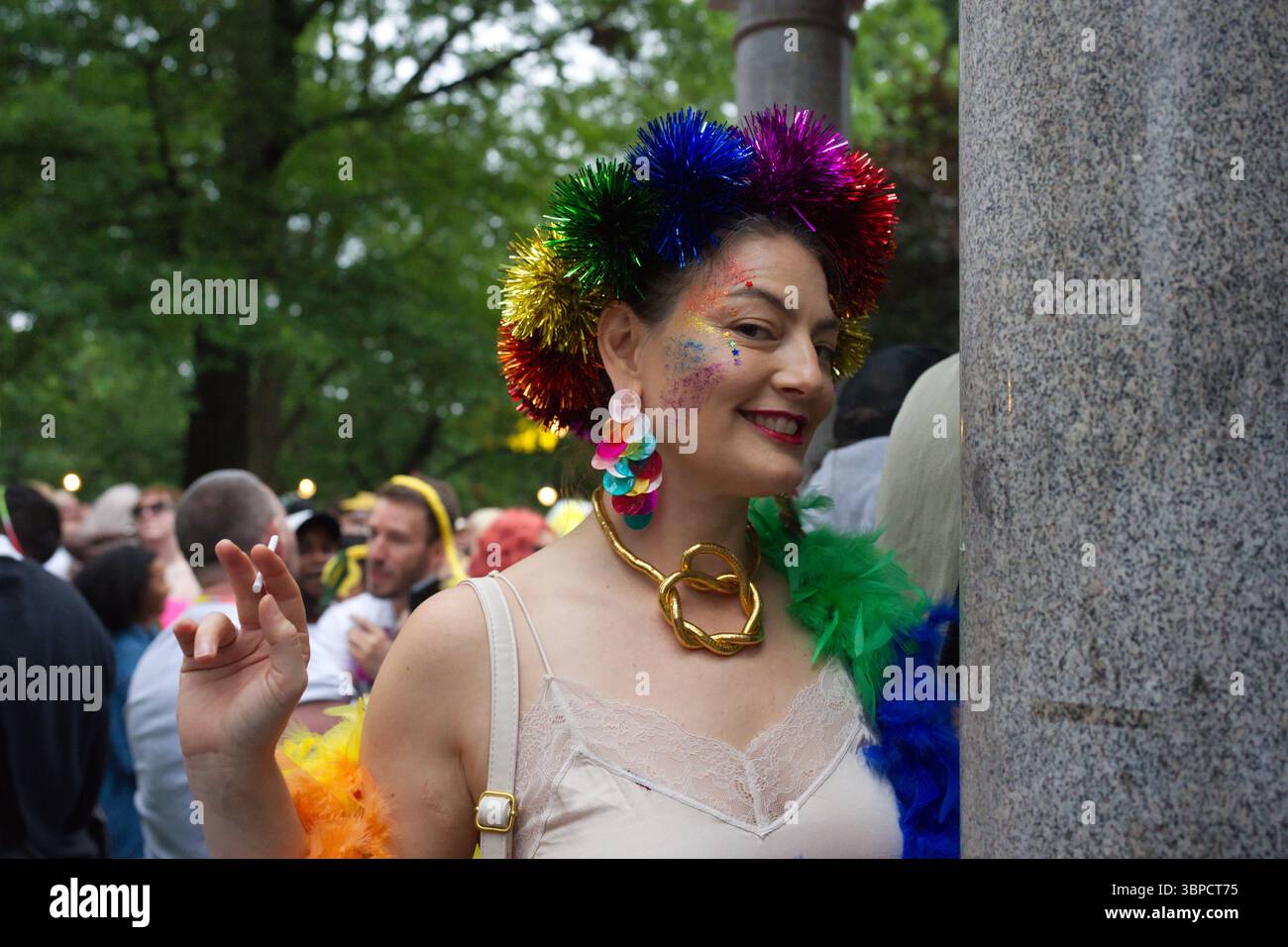 Une femme avec un maquillage de paillettes couleur arc-en-ciel et une coiffe festive, tenant une cigarette, se tient près d'un pilier de pierre lors d'un événement en plein air. Elle porte Banque D'Images