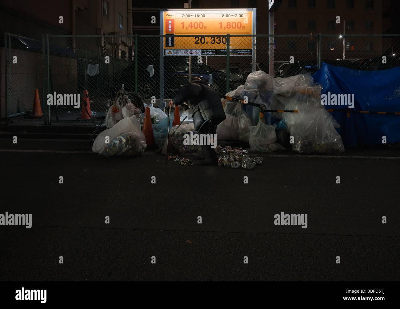 Homme âgé récupérant des canettes dans une rue de Tokyo la nuit, en utilisant un bâton pour ramasser les matières recyclables d'une pile de sacs poubelle Banque D'Images