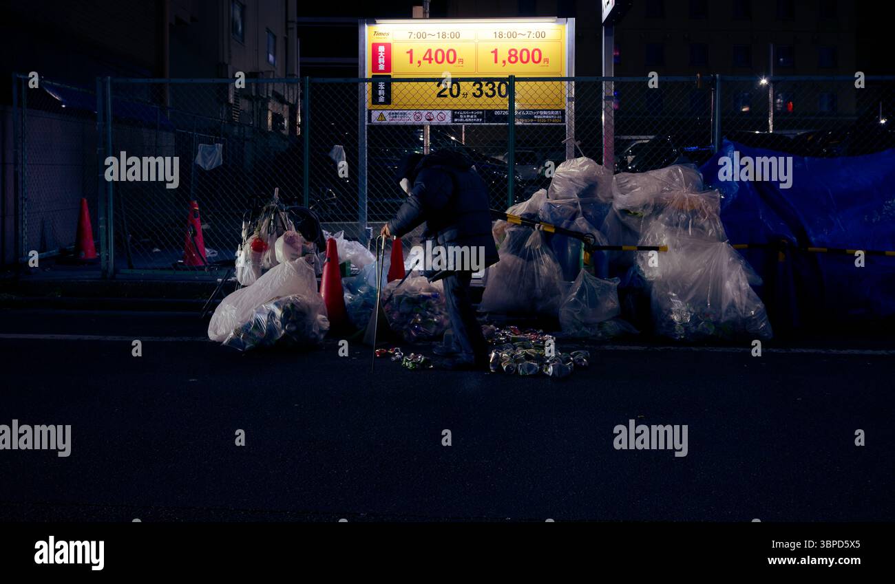 Un homme âgé récupère des boîtes de conserve avec un bâton de préhension parmi des tas d'ordures dans une rue sombre de Tokyo, sous la lueur d'un panneau de stationnement Banque D'Images