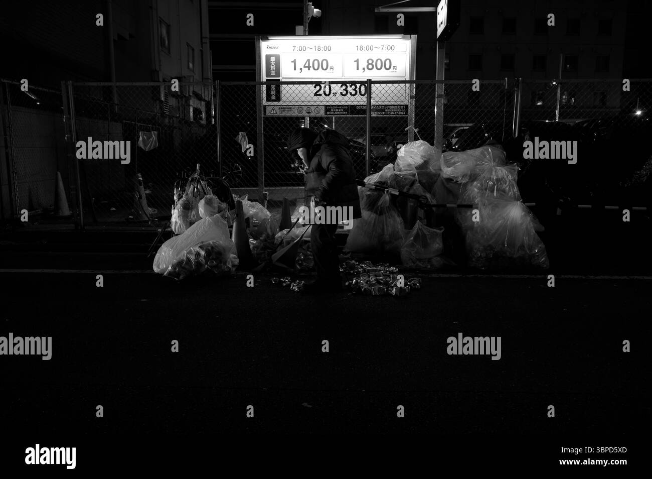 Un homme âgé trie des canettes avec une pince parmi les sacs poubelle dans une rue de Tokyo la nuit, illuminée par un panneau de parking Banque D'Images