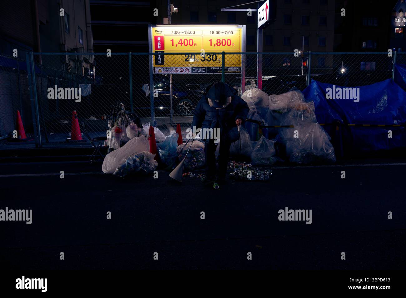 Un homme âgé récupère des boîtes de conserve avec un bâton de préhension parmi des tas d'ordures dans une rue sombre de Tokyo, sous la lueur d'un panneau de stationnement Banque D'Images