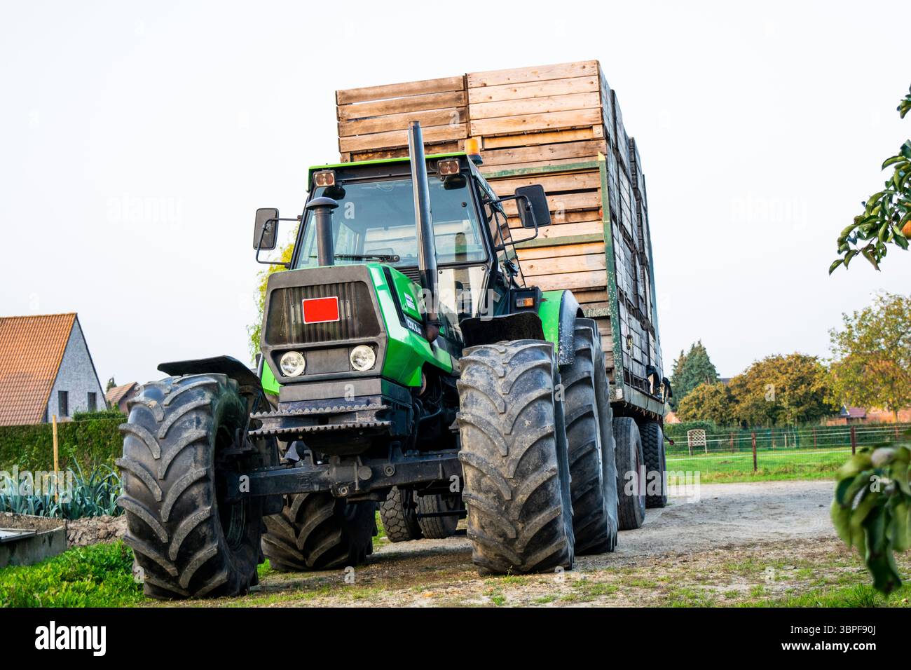 Grand tracteur agricole vert avec une remorque chargée de caisses en bois, garé sur un chemin de terre dans un cadre rural avec des bâtiments agricoles et des arbres. Banque D'Images