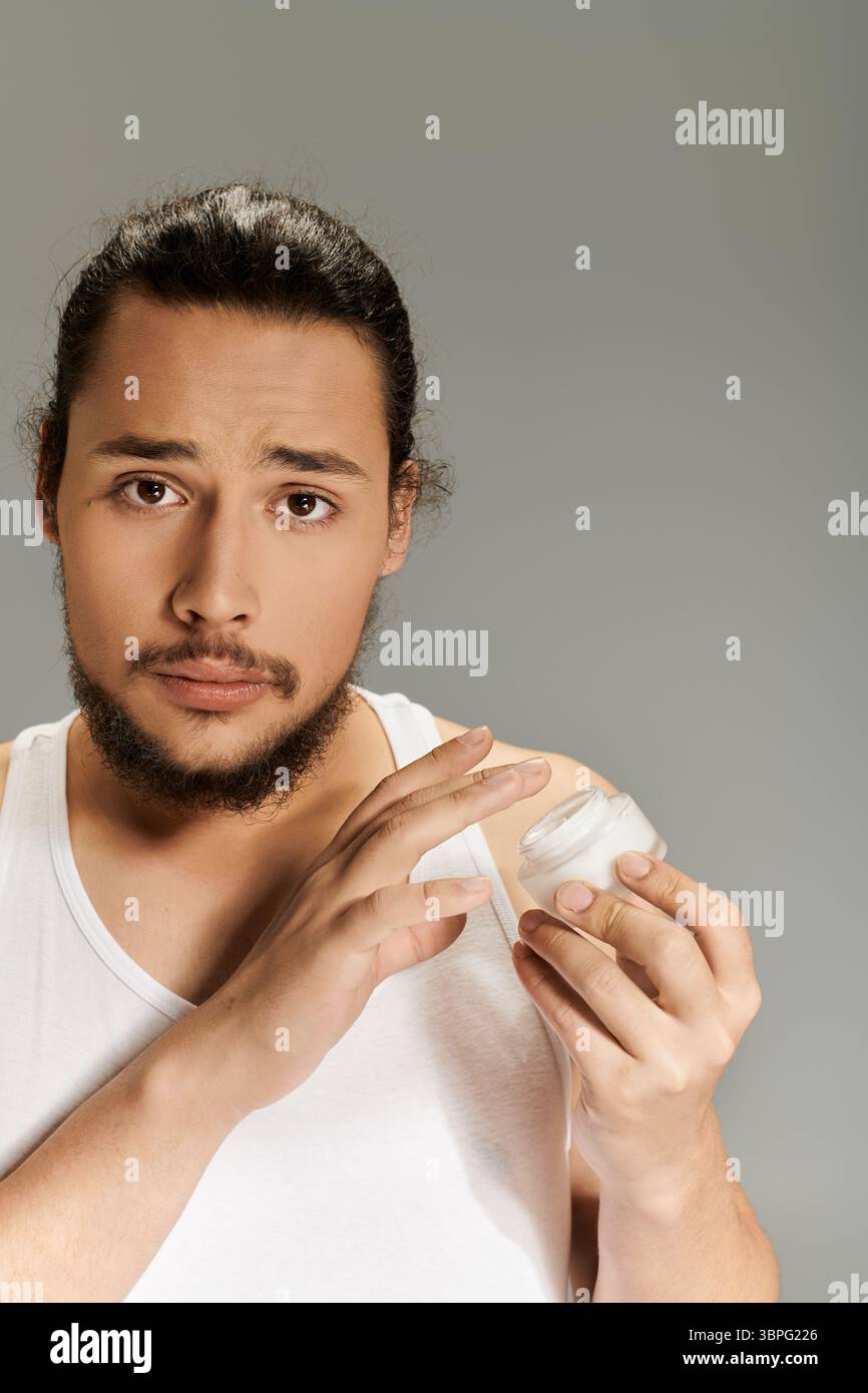 Jeune homme barbu tient le récipient de crème, se concentrant sur la routine de soins de la peau dans un environnement simple. Banque D'Images