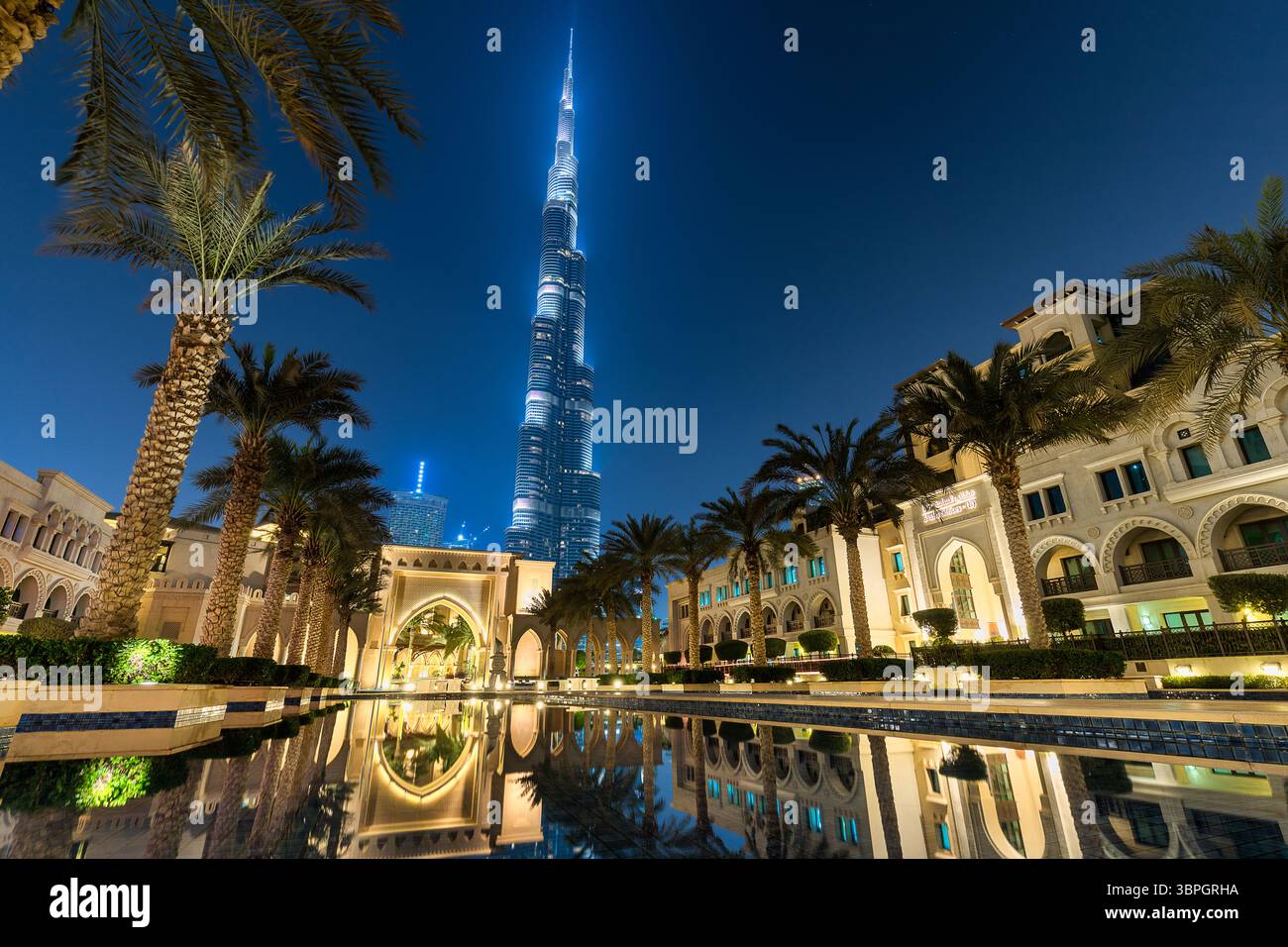 Vue de la Burj Khalifa illuminée surplombant l'architecture complexe reflétée dans l'eau sereine, encadrée par des palmiers, Dubaï, Dubaï, United Arab E Banque D'Images