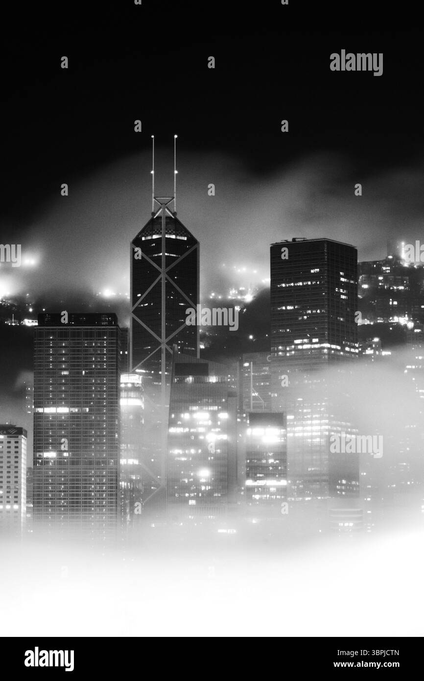 Une nuit brumeuse dans le quartier financier de Hong Kong capturée en noir et blanc, avec des gratte-ciel emblématiques entourés de brume et illuminés par des lumières. Banque D'Images