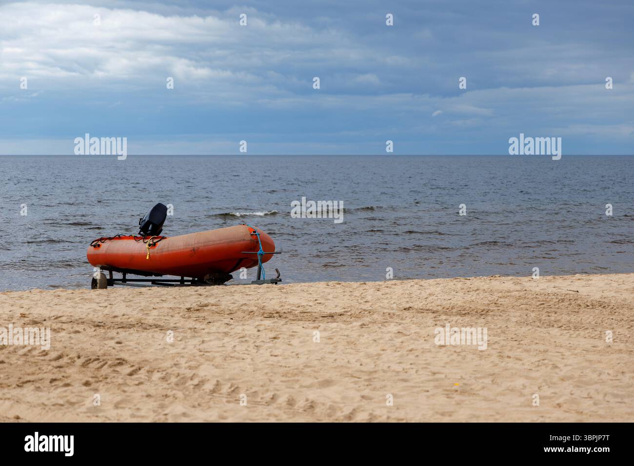 Un bateau gonflable avec un moteur est garé sur une remorque sur une rive sablonneuse, avec une mer calme en arrière-plan sous un ciel nuageux. Banque D'Images