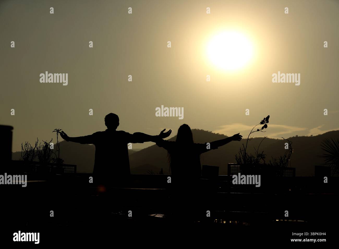 immortalisez la silhouette de deux personnes à bras ouvert face à un coucher de soleil ou à un lever de soleil. semblent être debout sur un toit avec la montagne en arrière-plan Banque D'Images