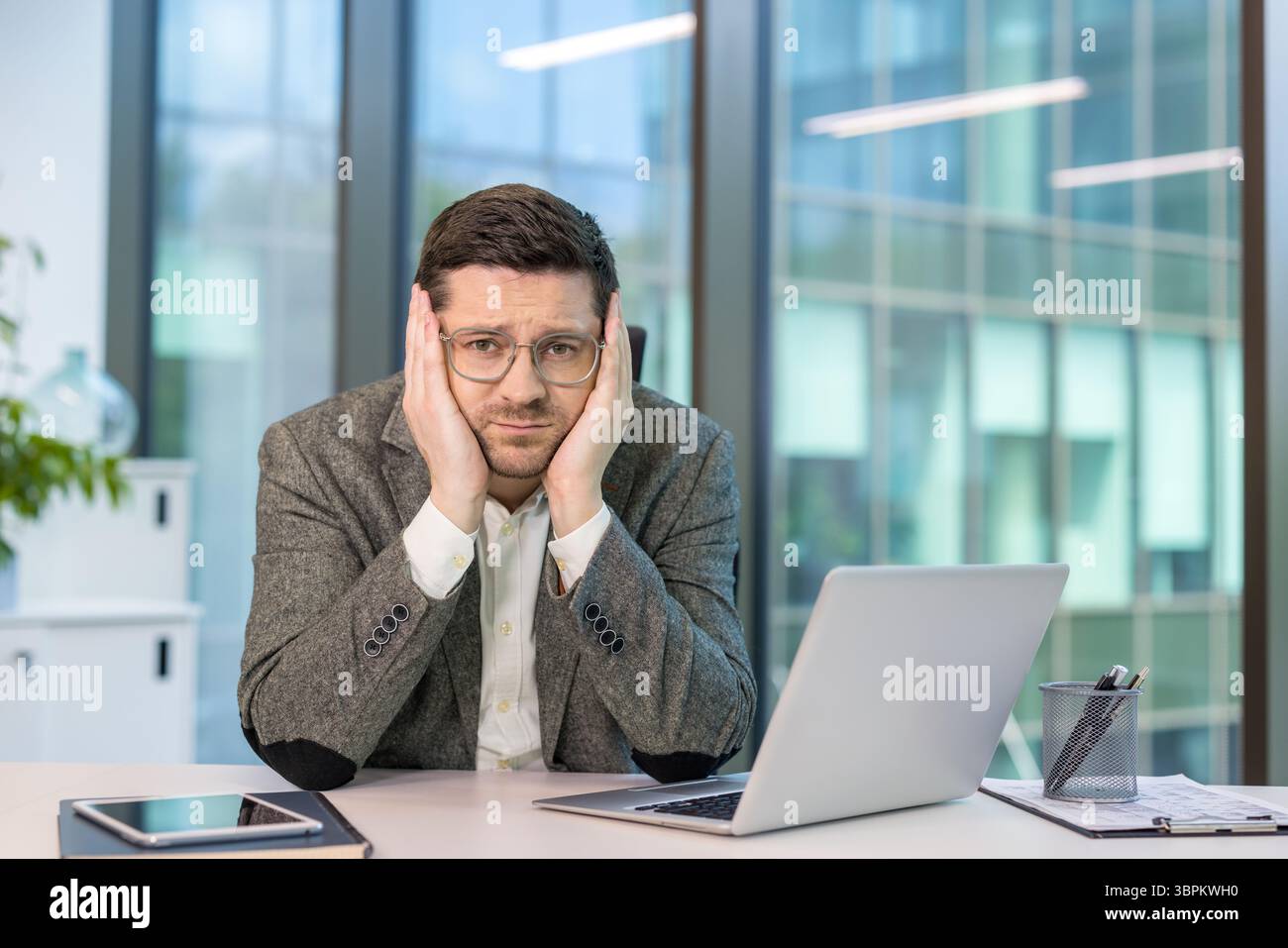 Portrait en gros plan d'un jeune employé de bureau assis à son bureau, la tête reposant sur ses mains, l'air ennuyé et fatigué devant la caméra. Banque D'Images