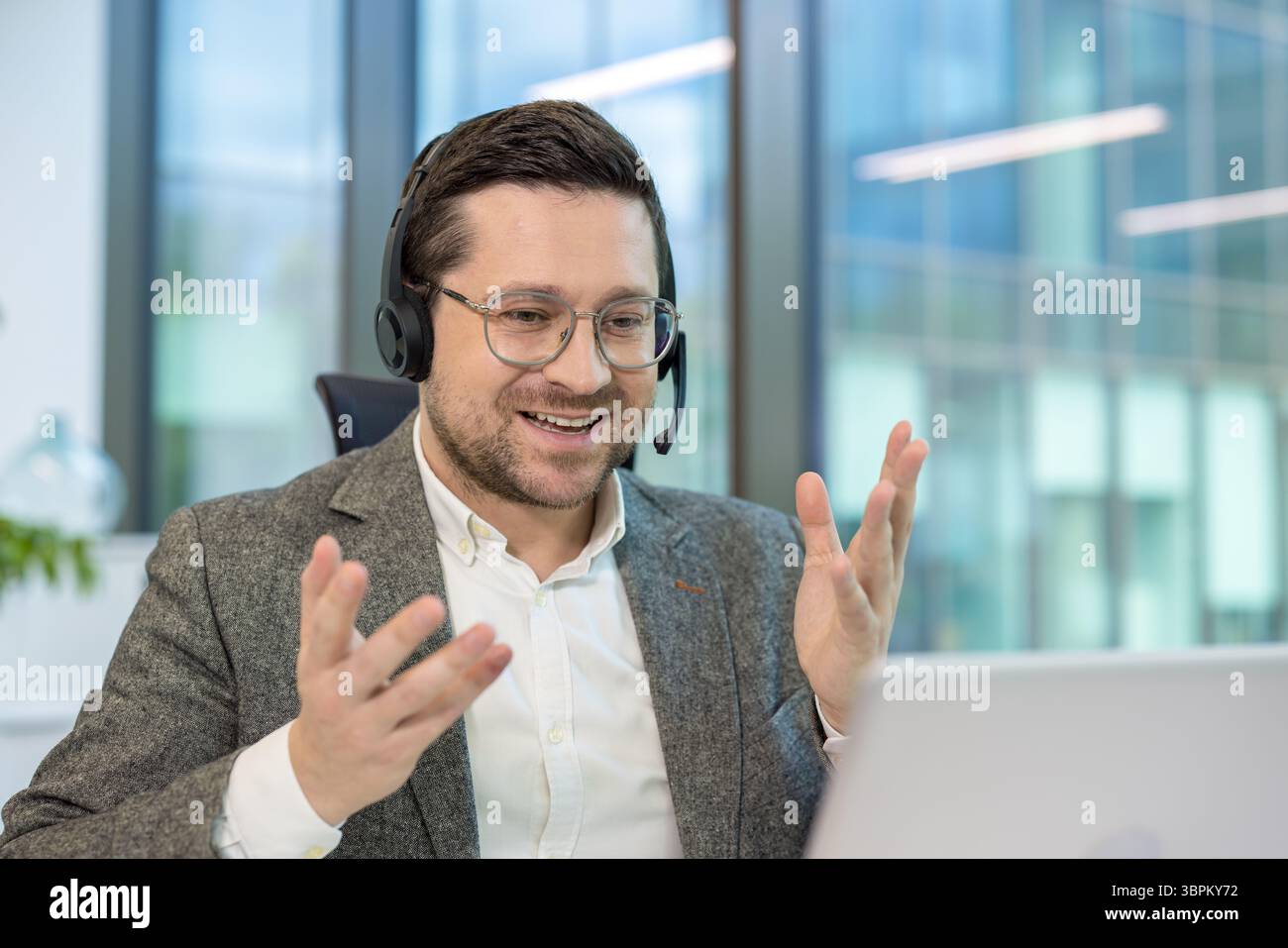 Un jeune homme de bureau est assis à un bureau dans le bureau, portant un casque et parlant lors d'un appel vidéo sur un ordinateur portable, faisant des gestes avec ses mains. Banque D'Images