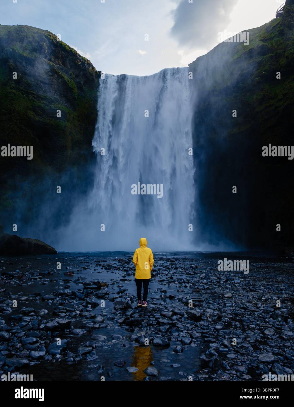 Découvrez la beauté époustouflante de la cascade de Skogafoss en Islande en tant que visiteur dans une veste de pluie jaune se tient fasciné par la chute d'eau pendant la lueur du coucher du soleil Banque D'Images