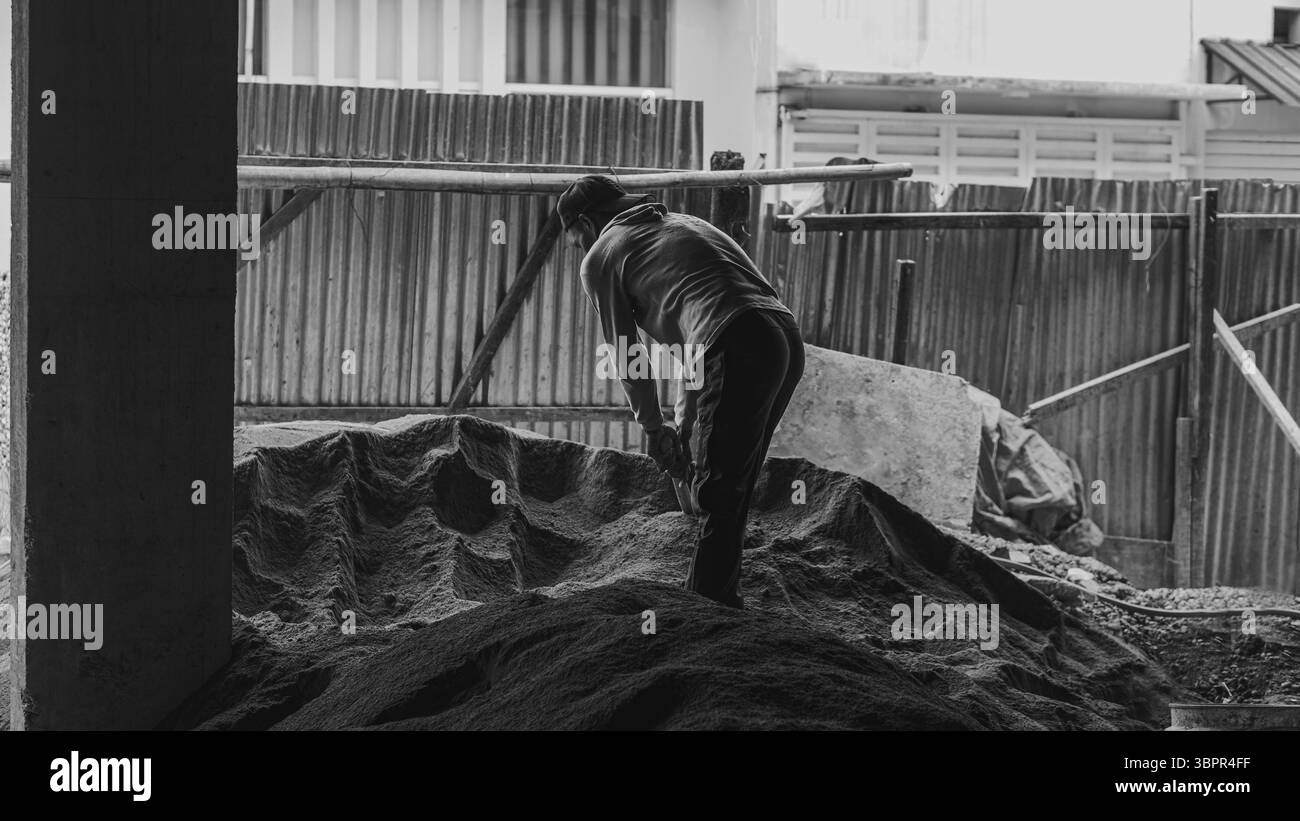 Un ouvrier de la construction houille manuellement du sable sur le site, montrant détermination, travail physique et persévérance dans la préparation du sol. Banque D'Images