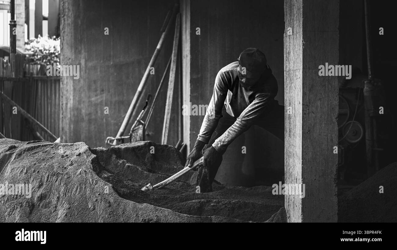 Un ouvrier de la construction houille manuellement du sable sur le site, montrant détermination, travail physique et persévérance dans la préparation du sol. Banque D'Images