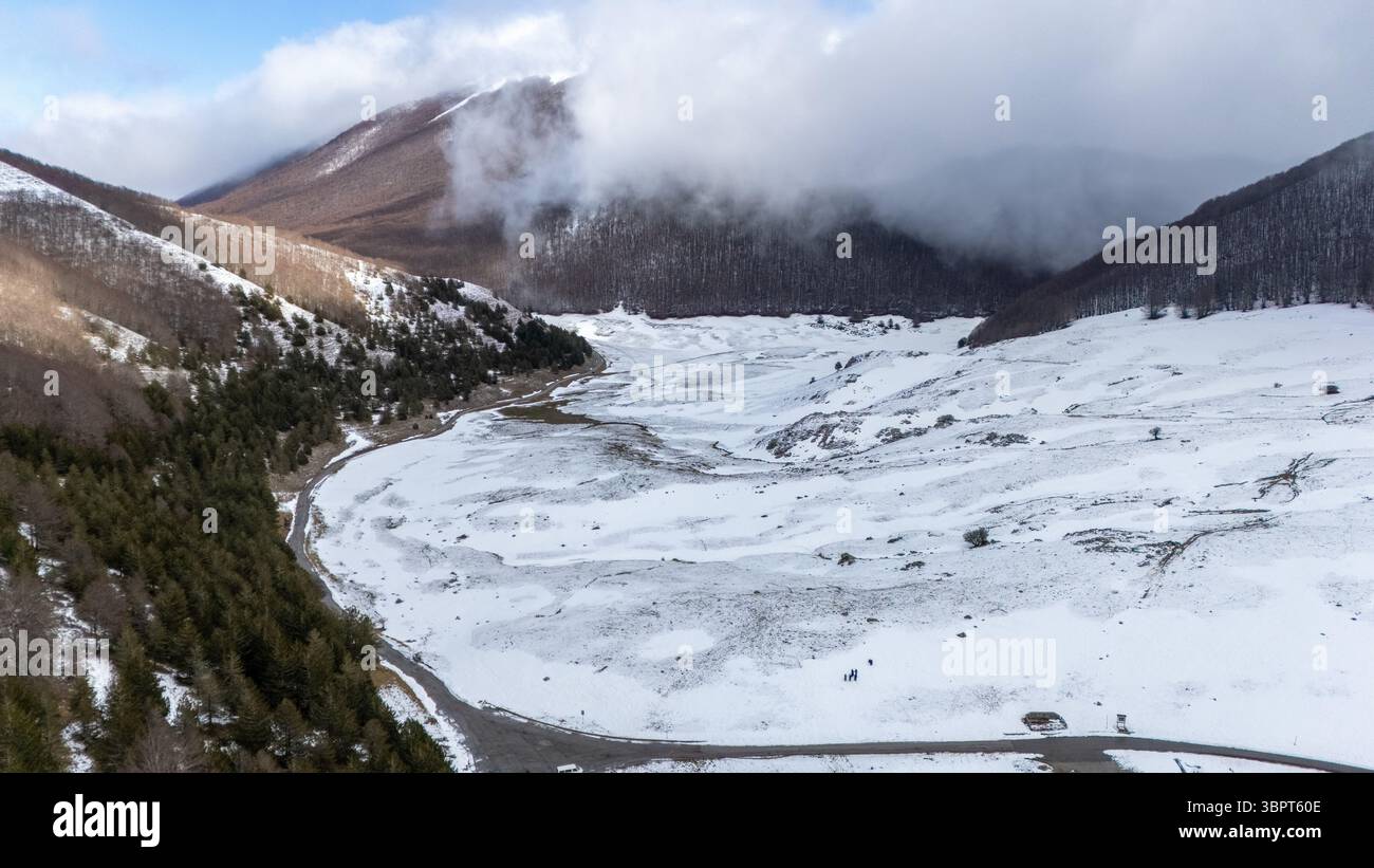 Vue aérienne d'une vallée enneigée entourée de montagnes et de forêts, avec des nuages couvrant partiellement les sommets. Le paysage est serein et intact Banque D'Images