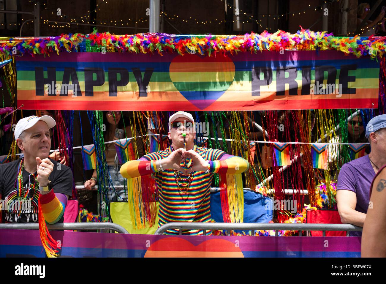 Une scène vibrante de défilé Pride avec un homme dans une chemise rayée arc-en-ciel soufflant un sifflet, donnant une forme de coeur avec ses mains. Une bannière « Happy Pride » Banque D'Images