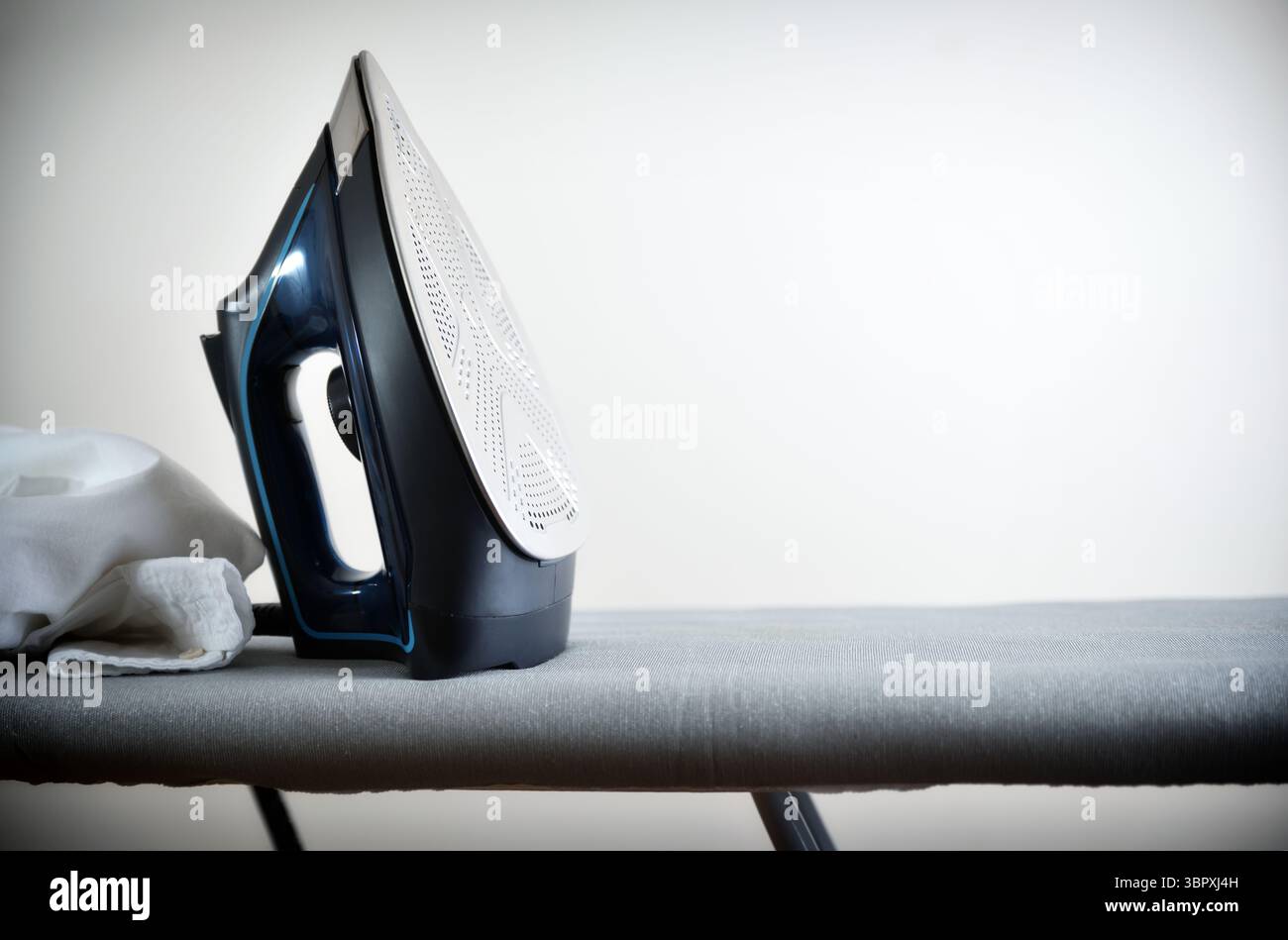 Concept de repassage de vêtements avec fer reposant verticalement sur une planche à repasser grise avec un vêtement blanc et un fond isolé léger. Vue de face. Banque D'Images