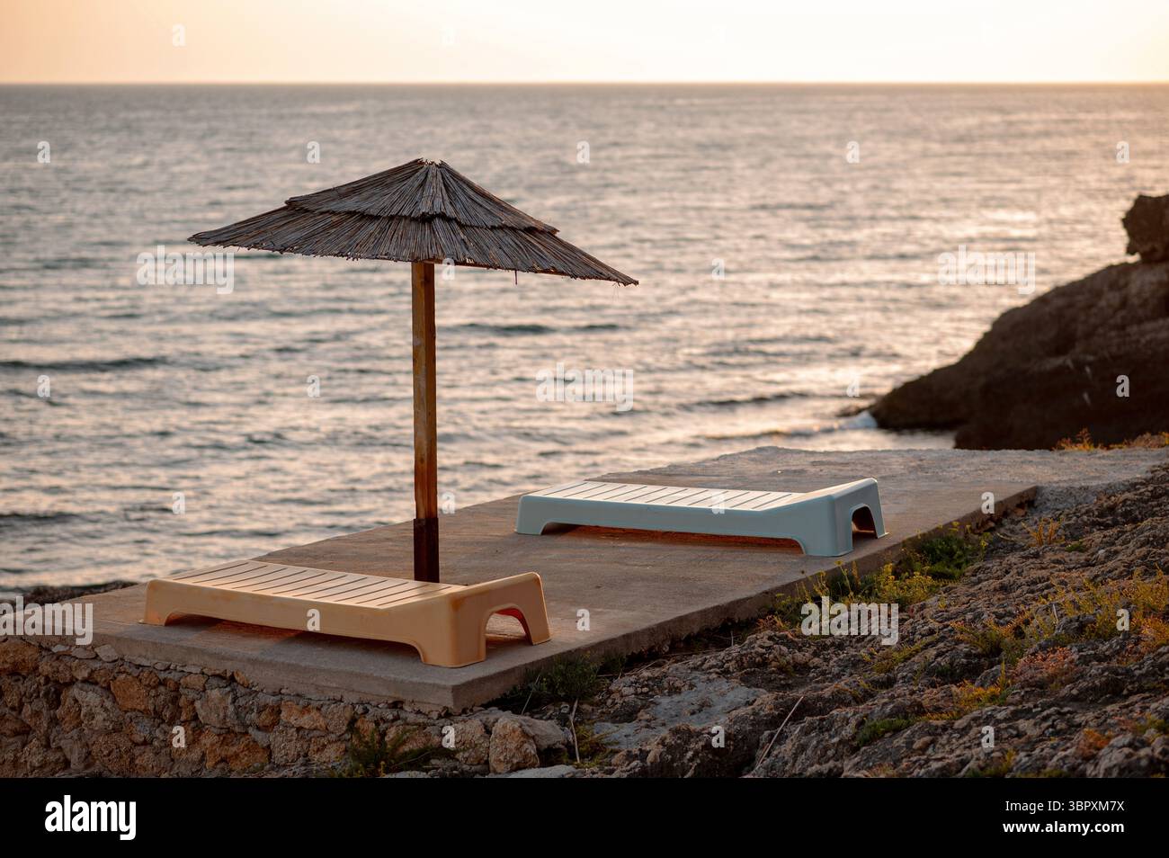 Deux chaises longues vides sous un parasol de paille sur une plage rocheuse au coucher du soleil. Banque D'Images