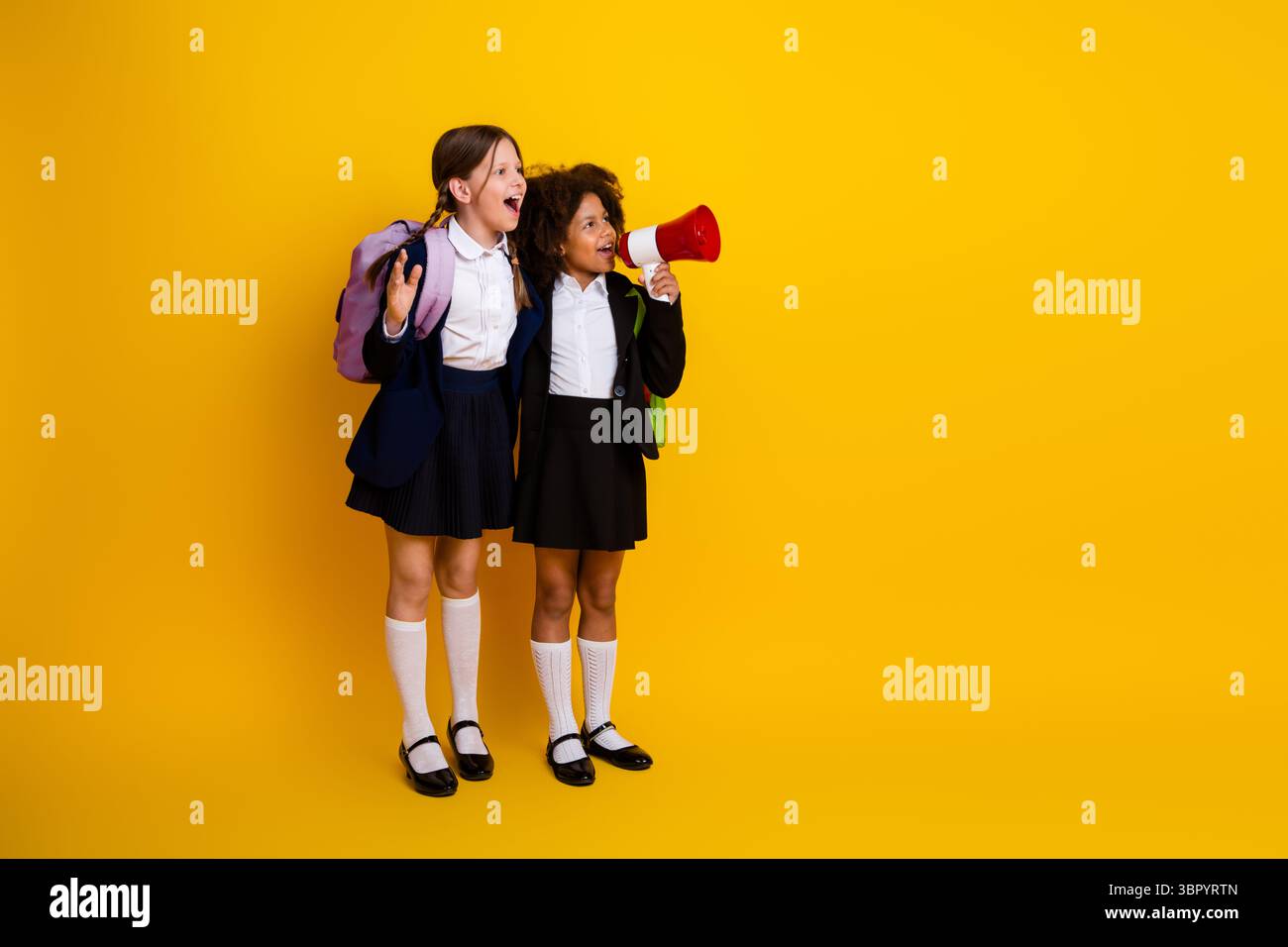 Deux écolières joyeuses portant des uniformes debout ensemble sur un fond jaune, exprimant leur amitié tout en tenant un mégaphone Banque D'Images