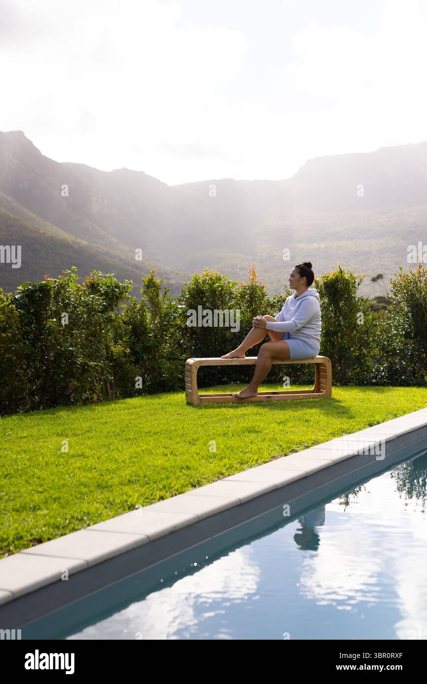 Femme asiatique adulte moyenne assise sur un banc en bois à côté d'une piscine rectangulaire sur une pelouse luxuriante, espace de copie. Tranquille, loisirs, nature, paysage, bien-être, outdoo Banque D'Images