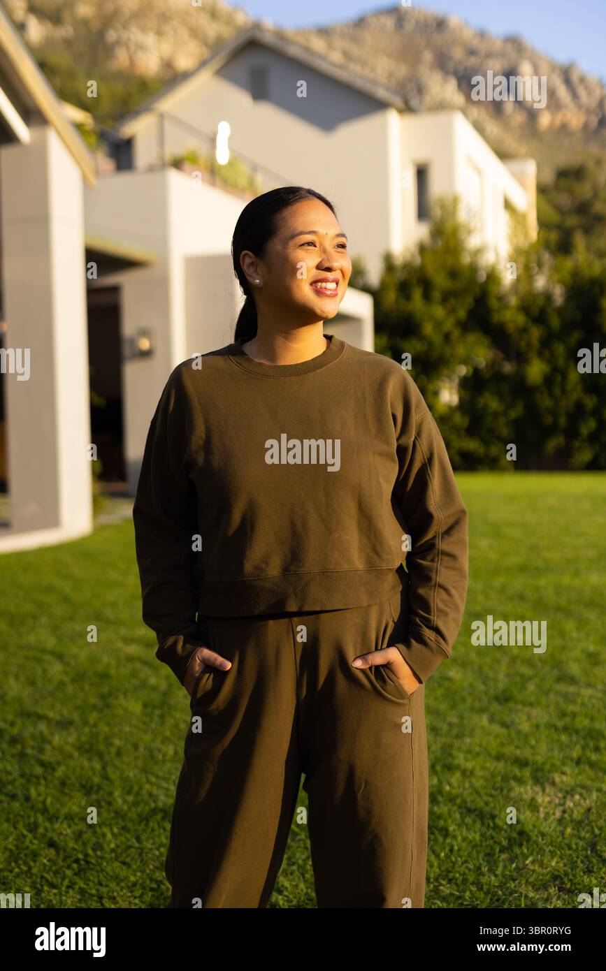 Femme asiatique adulte moyenne souriant sur la pelouse d'herbe à la maison blanche portant une tenue verte décontractée. Extérieur, décontracté, minimaliste, style de vie, naturel, pittoresque, serein Banque D'Images Femme asiatique adulte moyenne souriant sur la pelouse d'herbe à la maison blanche portant une tenue verte décontractée. Extérieur, décontracté, minimaliste, style de vie, naturel, pittoresque, serein Banque D'Images