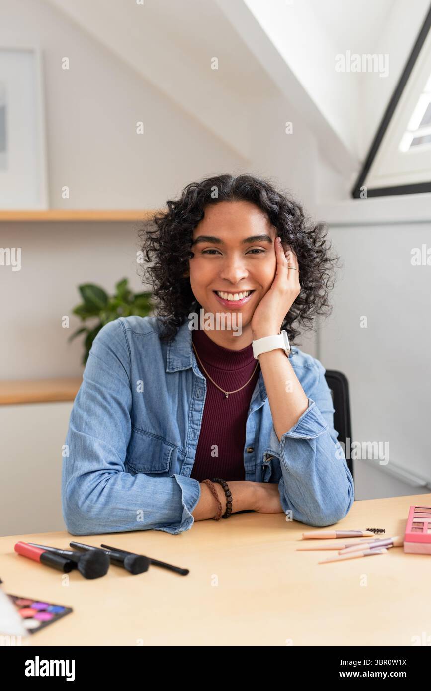 Personne non binaire souriant tout en se reposant la tête sur la main au bureau du grenier avec palette de maquillage et pinceaux. Créatif, cosy, minimaliste, artistique, inspirant, m Banque D'Images