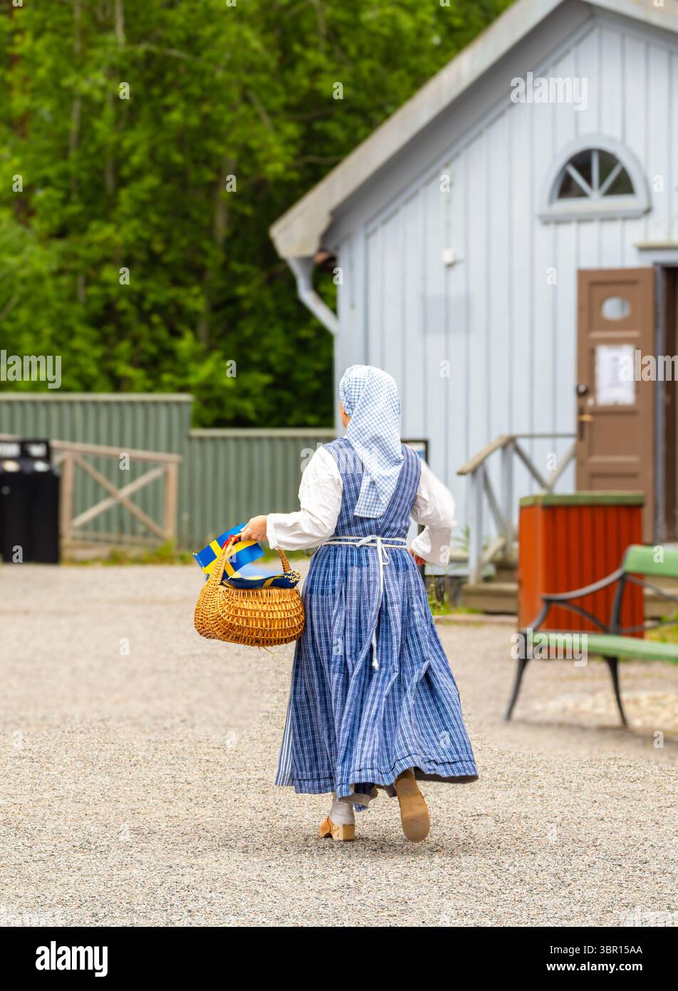 Sodertalje, Suède - 6 juin 2025 : une suédoise dans une robe traditionnelle bleue et blanche portant un panier tissé, s'éloignant d'un bâtiment dans un RU Banque D'Images
