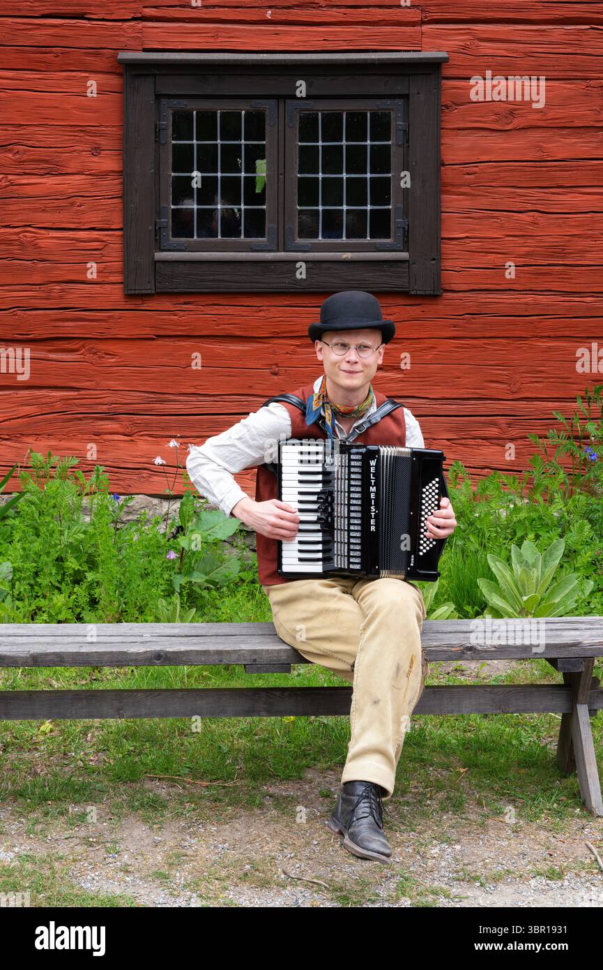 Sodertalje, Suède - 6 juin 2025 - Un homme jouant de l'accordéon assis sur un banc devant un bâtiment rustique en bois rouge. Banque D'Images