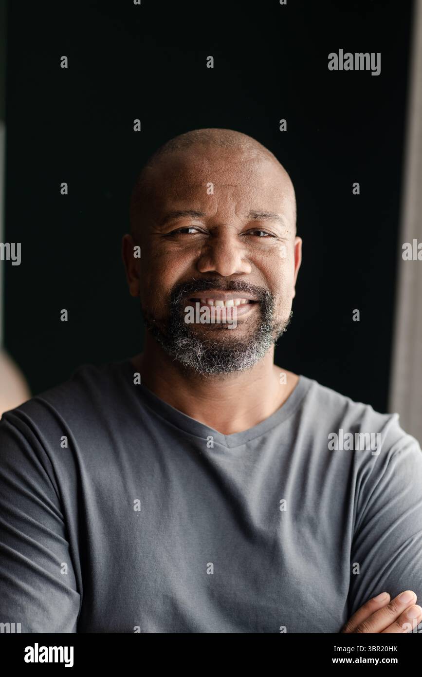 Homme afro-américain senior debout avec les bras croisés dans le studio près de la fenêtre portant une chemise grise. Portrait, confiance, minimalisme, introspection, natu Banque D'Images